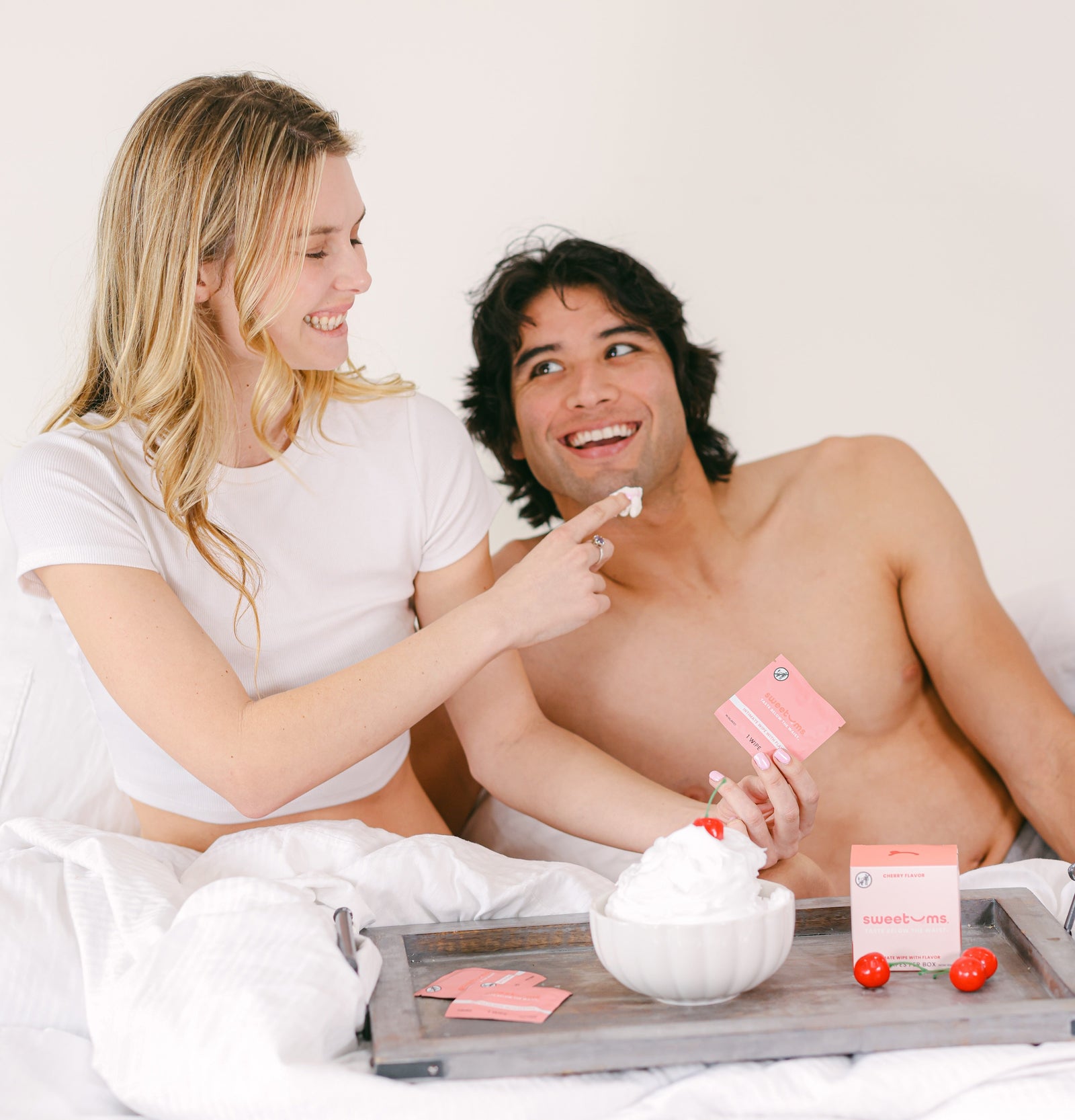 woman holding cherry sweetums wipes in bed with a man and giving him a taste of whipped cream on her finger