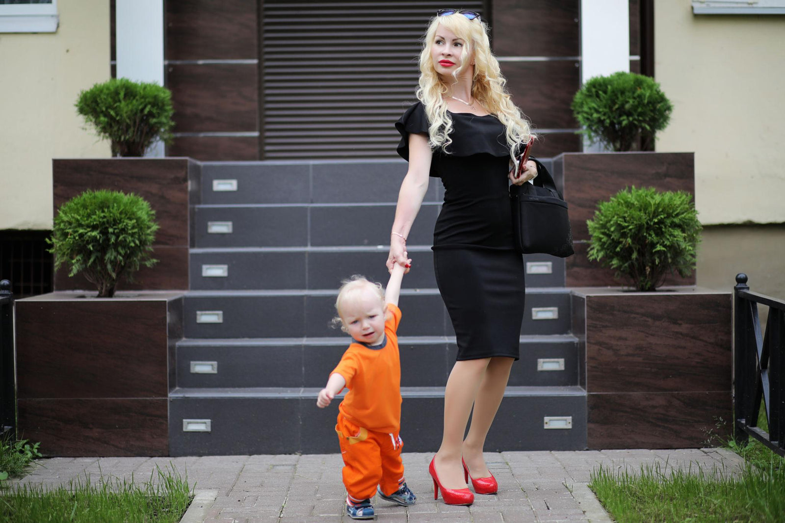woman in heels and a dress holding the hand of a toddler in front of a building