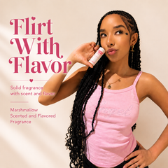 A woman with long black braids wears a light pink tank top and holds a Sweet Spot Marshmallow fragrance near her face. Large pink text on the left reads 'Flirt With Flavor' with smaller text describing the fragrance as marshmallow-scented and flavored.
